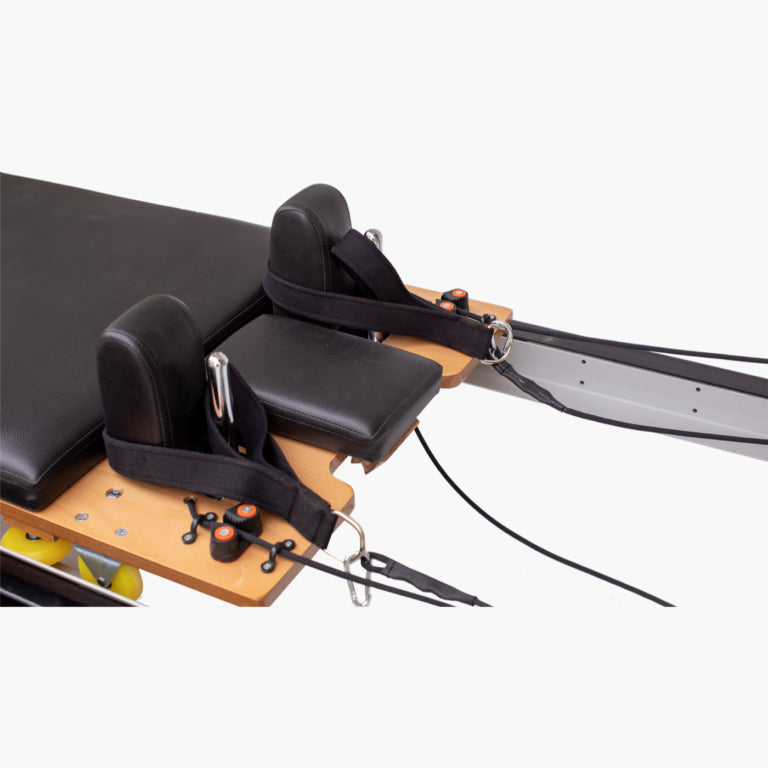 Set Pilates Home Metal Reformer machine close-up showing black padded headrests, black straps, wooden carriage, metal guide bars, and resistance cords on a light gray background.