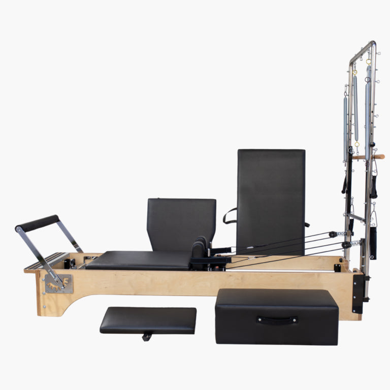 SET Pilates Master Wood Reformer with Tower with black padded mats and straps, shown on a plain light gray background. A separate black foot platform and a black storage box are placed in front of the machine.