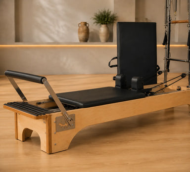 A wooden Pilates reformer machine with black padded foam elements, shown in a minimalist studio setting with light wood floors and a warm beige background.