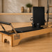 A wooden Pilates reformer machine with black padded foam elements, shown in a minimalist studio setting with light wood floors and a warm beige background.