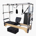 SET Pilates SECR Wooden Cadillac Reformer Trapeze Table with wooden frame, black padded carriage and seat, hanging resistance bands, and a separate black foam block on a plain light gray background.