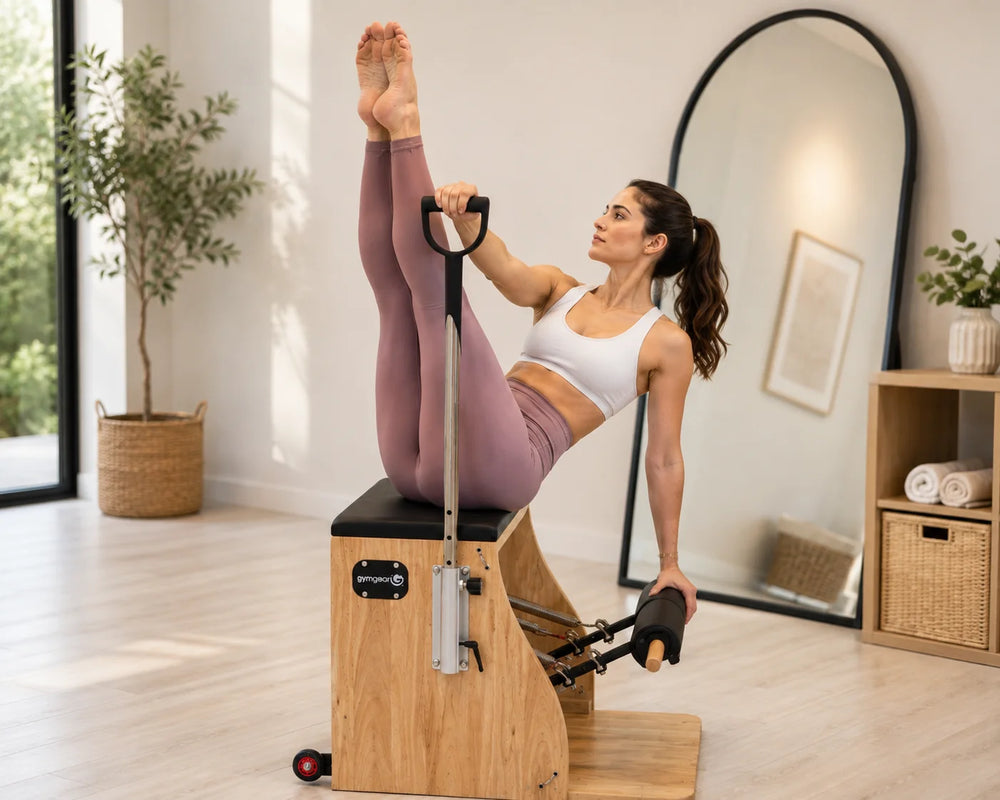 A woman in a white sports bra and mauve leggings performs a Pilates exercise on a wooden reformer machine in a bright, modern home gym.