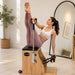 A woman in a white sports bra and mauve leggings performs a Pilates exercise on a wooden reformer machine in a bright, modern home gym.