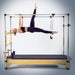 A woman in black activewear performs a yoga pose suspended from a wooden and metal Pilates reformer machine against a light gray studio background.