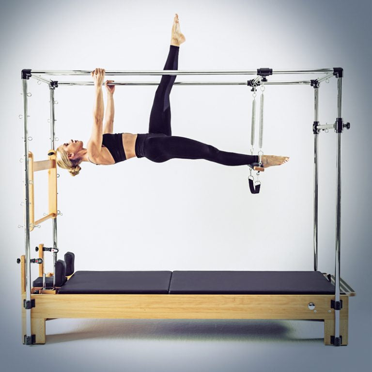 A woman in black activewear performs a yoga pose suspended from a wooden and metal Pilates reformer machine against a light gray studio background.