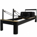 Origin Fitness Pilates reformer machine with a black frame, natural wood rails, and black straps, shown on a solid white background.