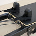 Close-up of a Pilates reformer machine showing the black padded footboard, black straps, and light wood edge trim on a beige floor.