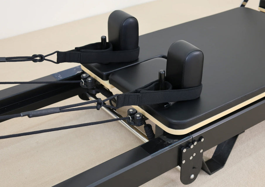 Close-up of a Pilates reformer machine showing the black padded footboard, black straps, and light wood edge trim on a beige floor.
