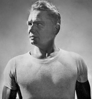 Black and white portrait of a Joseph Pilates wearing a fitted short-sleeve crew-neck t-shirt, looking off to the side.