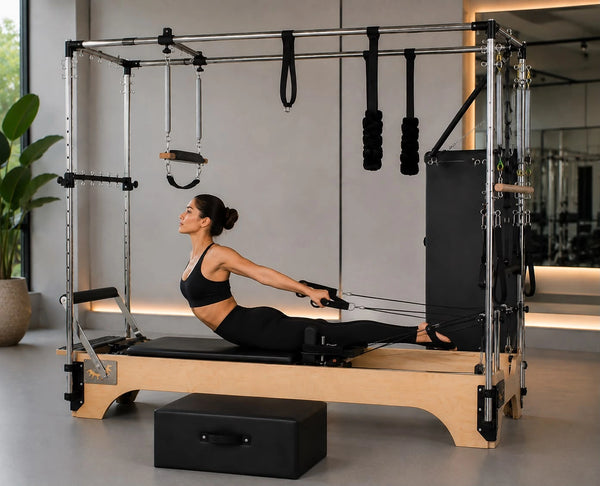 A woman in a black sports bra and leggings performs a Pilates exercise on a wooden and metal reformer machine in a bright, modern gym with a large window and potted plants.