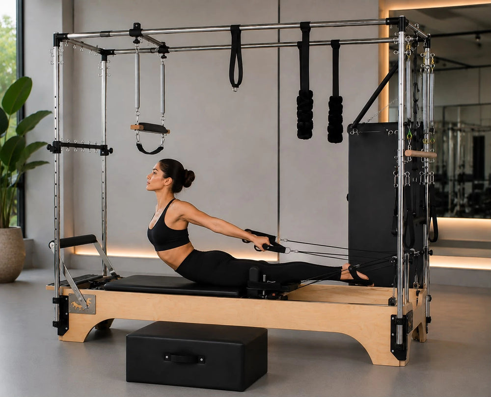A woman in a black sports bra and leggings performs a Pilates exercise on a wooden and metal reformer machine in a bright, modern gym with a large window and potted plants.