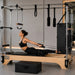 A woman in a black sports bra and leggings performs a Pilates exercise on a wooden and metal reformer machine in a bright, modern gym with a large window and potted plants.