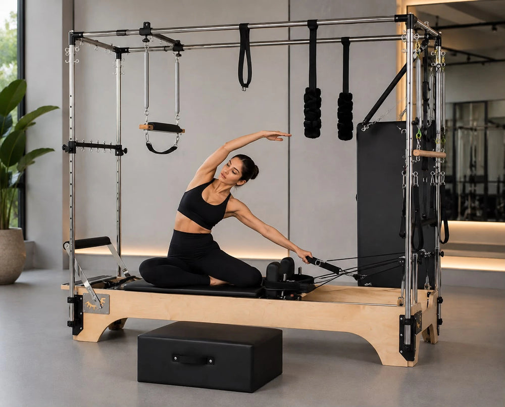 A woman in a black sports bra and leggings performs a side stretch on a wooden Pilates reformer machine with metal frame bars, black resistance bands, and a black foam block in a bright modern studio.