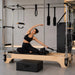 A woman in a black sports bra and leggings performs a side stretch on a wooden Pilates reformer machine with metal frame bars, black resistance bands, and a black foam block in a bright modern studio.