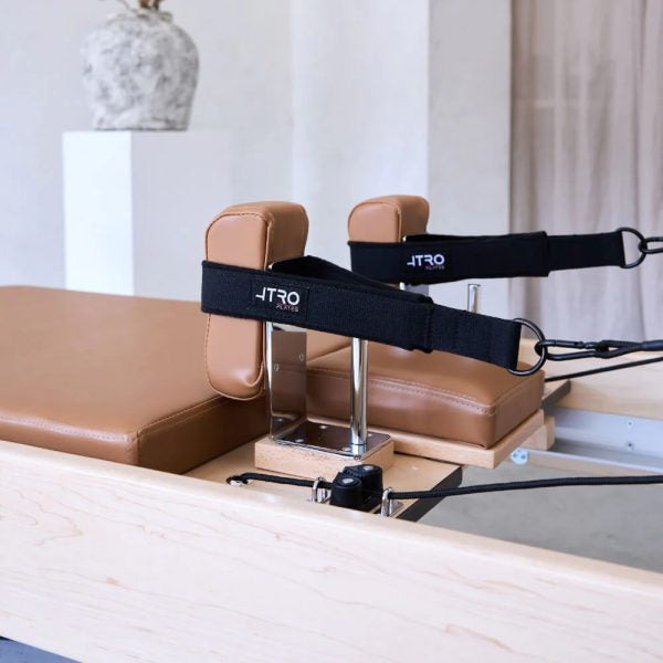 A Pilates reformer machine with tan leather padding, a light wood frame, and black straps branded