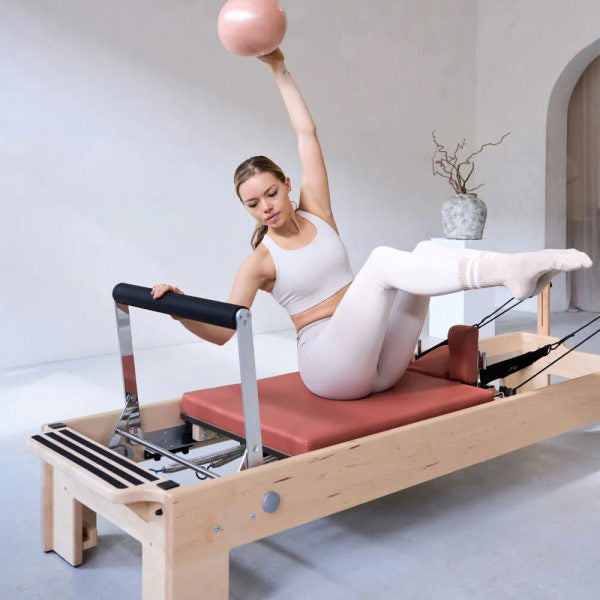 A woman  sits on a wooden Pilates reformer machine, holding a pink exercise ball overhead with one arm while extending the other leg. The room has a light gray wall and floor, with a small vase on a side table in the background.