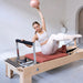 A woman  sits on a wooden Pilates reformer machine, holding a pink exercise ball overhead with one arm while extending the other leg. The room has a light gray wall and floor, with a small vase on a side table in the background.