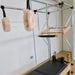 Pilates reformer machine with a wooden frame, black padded carriage, metal support bars, and two beige faux-fur handles hanging from a silver pull-up bar frame in a studio room.