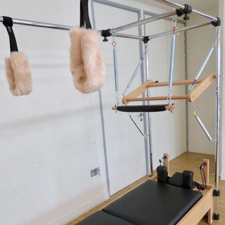 Pilates reformer machine with a wooden frame, black padded carriage, metal support bars, and two beige faux-fur handles hanging from a silver pull-up bar frame in a studio room.