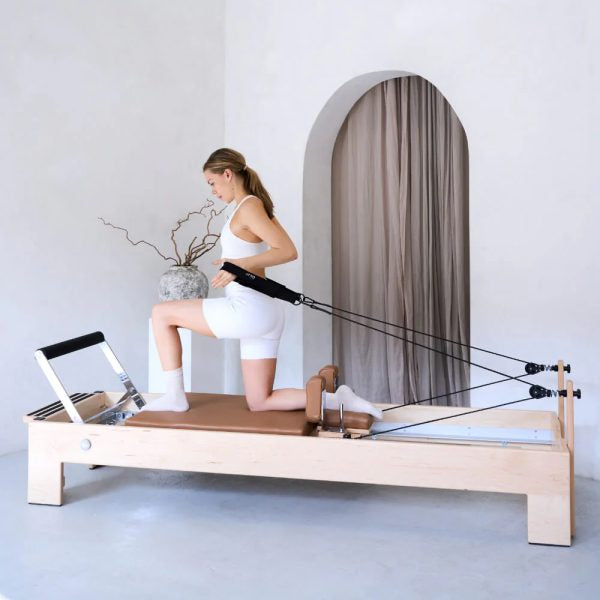 A woman kneels on a wooden Pilates reformer machine, using resistance bands. The setting is a minimalist studio with a light gray floor, a white wall, and an arched doorway with a brown curtain.