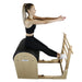 A woman sits in a wooden reformer Pilates machine with a black padded seat, shown against a plain white background.