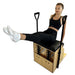 A woman sits on a wooden Pilates reformer machine with a black padded seat, demonstrating a leg extension exercise. She is wearing white socks, and the background is solid white.