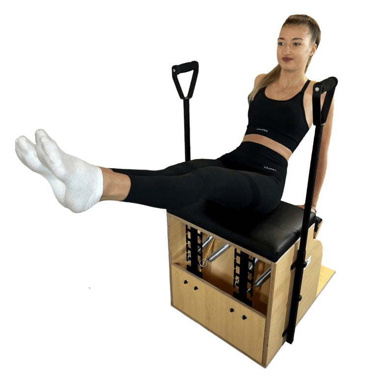 A woman sits on a wooden Pilates reformer machine with a black padded seat, demonstrating a leg extension exercise. She is wearing white socks, and the background is solid white.