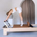 A woman performs a forward bend exercise on a wooden Pilates reformer machine in a minimalist studio with beige walls and a gray curtain.