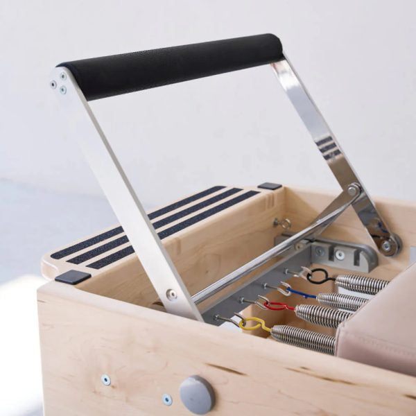 Close-up of a Pilates reformer machine showing the wooden frame, black foam handle, metal support arms, springs, and adjustment knobs.