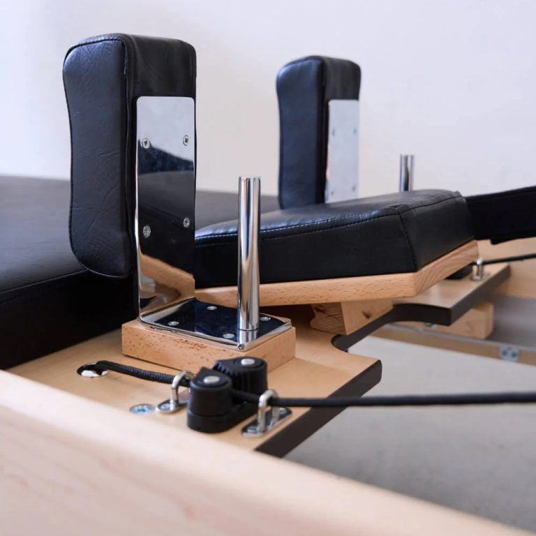 Pilates reformer machine close-up showing black padded handles, wooden frame, metal posts, and black resistance cords on a light wood platform.