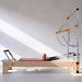 A wooden Pilates reformer machine with a pinkish-red cushion, metal springs, and a silver support frame, photographed in a minimalist studio with diagonal sunlight on a white wall.