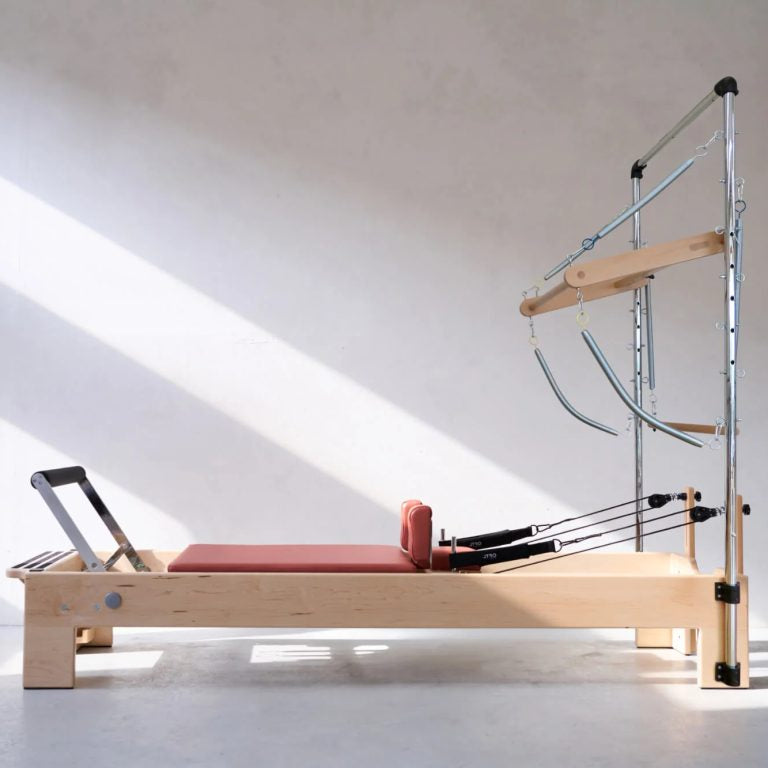 A wooden Pilates reformer machine with a pinkish-red cushion, metal springs, and a silver support frame, photographed in a minimalist studio with diagonal sunlight on a white wall.