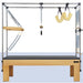 A Pilates reformer machine with a natural wood frame, black padded platform, metal tension rods, and hanging yellow resistance loops, shown against a plain white background.