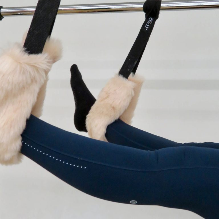 A close-up of a person's legs in navy blue leggings extended over a black suspension reformer machine with fluffy cream faux-fur cuffs.