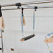 Pilates reformer machine components including a metal tension bar, silver spring attachments, a black foot bar, and a beige fabric strap hanging against a plain white wall.