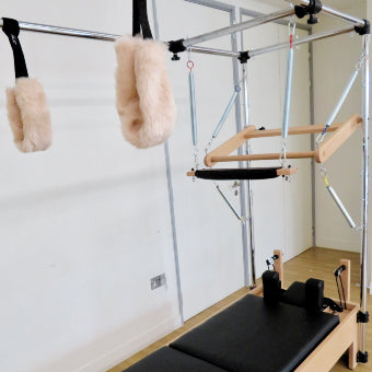 Pilates reformer machine with a wooden frame, black padded carriage, hanging straps, and fluffy cream grips in a bright indoor studio with white walls and wood floor.