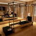 A Pilates reformer machine with a wooden frame, black carriage, and hanging rope attachments, photographed in a modern wood-paneled studio with mirrors and exercise equipment in the background.
