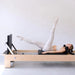 A woman in light beige activewear exercises on a wooden Pilates reformer machine with a black padded bed and black handlebar, set against a plain light gray studio background.
