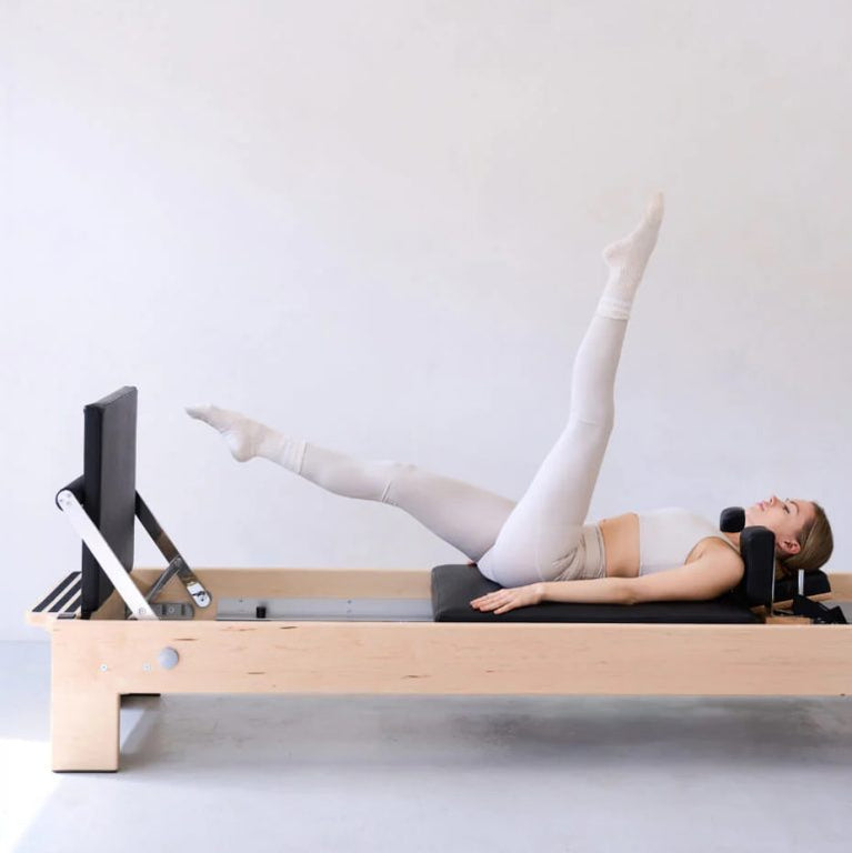 A woman in light beige activewear exercises on a wooden Pilates reformer machine with a black padded bed and black handlebar, set against a plain light gray studio background.
