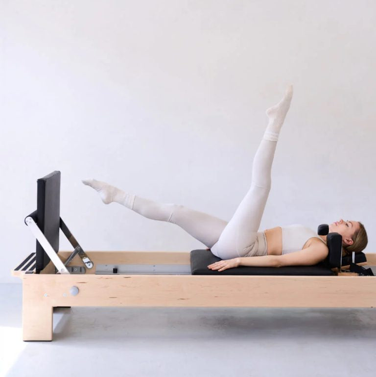 A woman in light beige activewear exercises on a wooden Pilates reformer machine with a black padded bed and black handlebar, set against a plain light gray studio background.