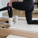 A person in black leggings performs a Pilates exercise on a wooden reformer machine with white padding in a bright studio.