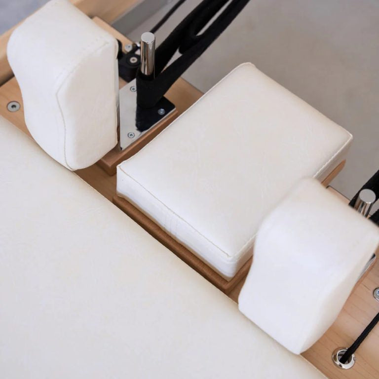 Close-up of a Pilates reformer machine with white padded footrest supports and wooden frame on a light neutral background.