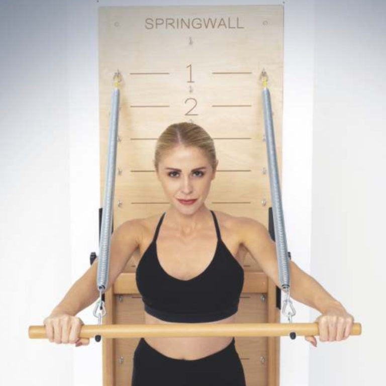 A woman holds a wooden reformer bar in front of a wooden Springwall exercise machine.