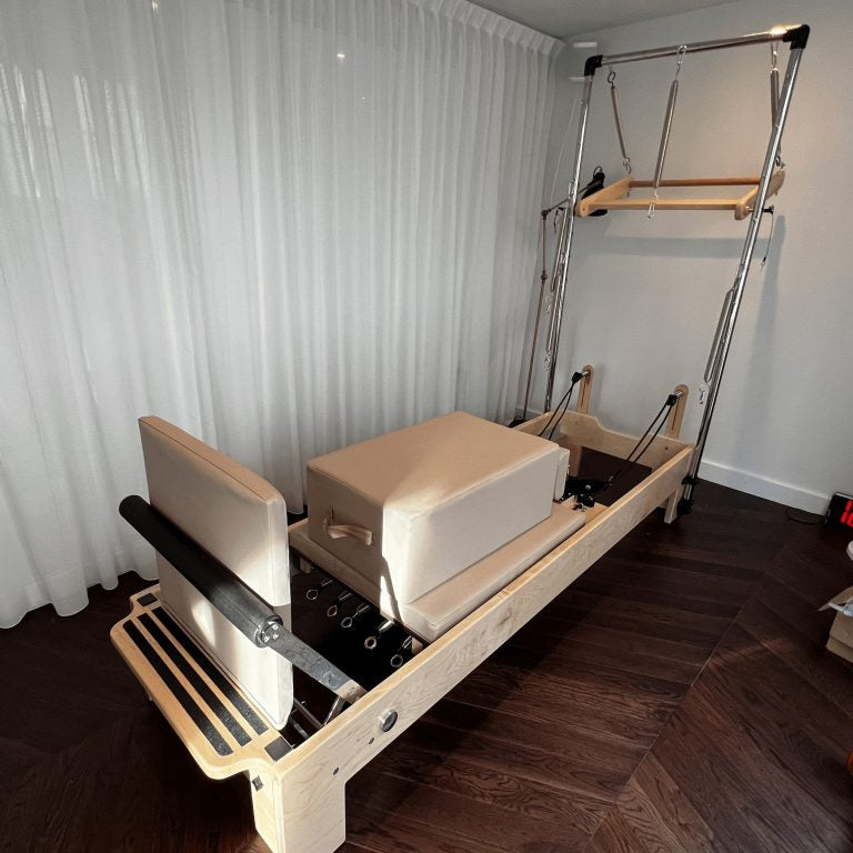 A wooden Pilates reformer machine with beige foam blocks and black straps, photographed indoors against white curtains and a white wall on dark wood flooring.