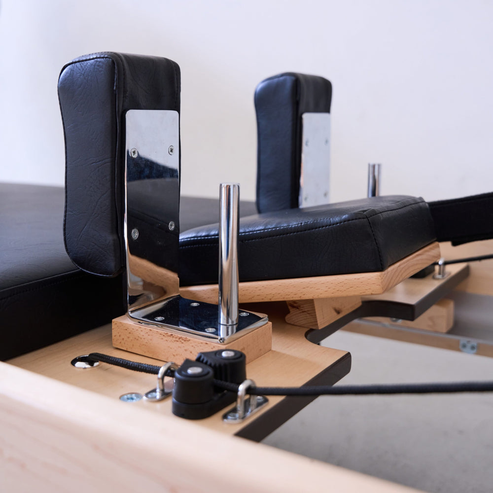 Pilates reformer machine with black padded straps, natural wood frame, and chrome metal guide rods on a light gray studio floor.