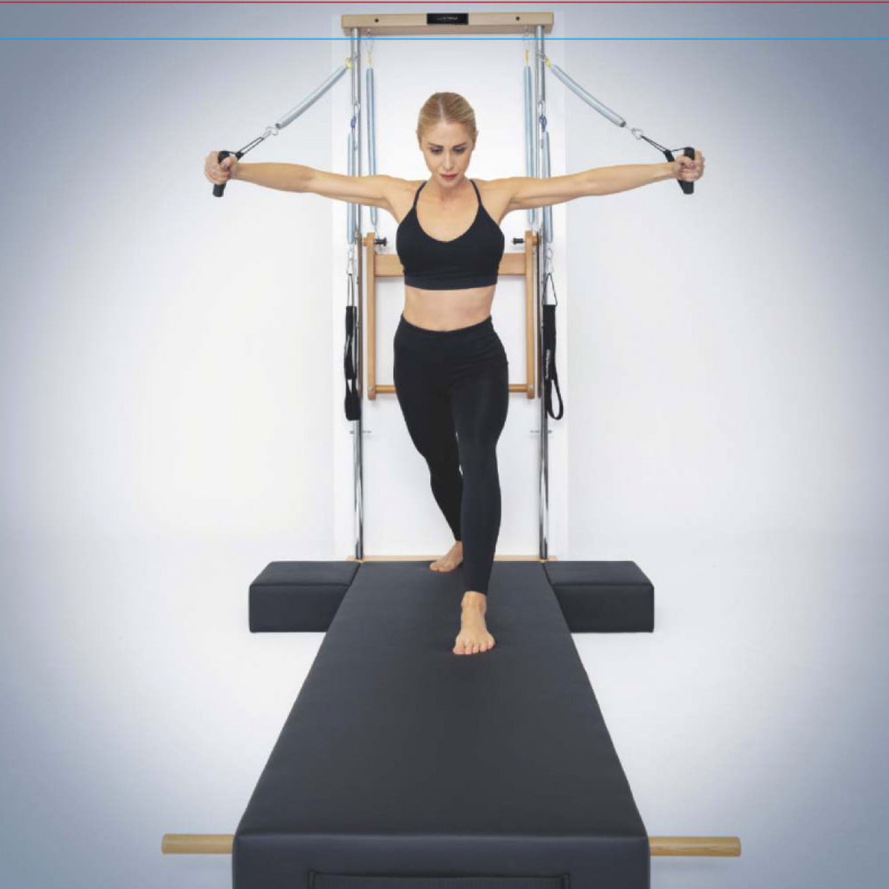 A woman on Atro Pilates Wall Tower Unit with Premium Mat holding handles with both arms extended. The reformer has a dark gray padded mat, wooden rails, and metal springs against a light gray studio background.