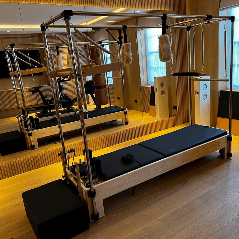 A Pilates reformer machine with a light wood frame, black padded carriage, and hanging straps, photographed inside a mirrored studio with wood-paneled walls and floor.