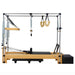 A Pilates reformer machine with a natural wood frame, black padded carriage, metal guide rails, spring-loaded resistance, hanging U-hand grips, and a black weight stack box, photographed on a plain white background.