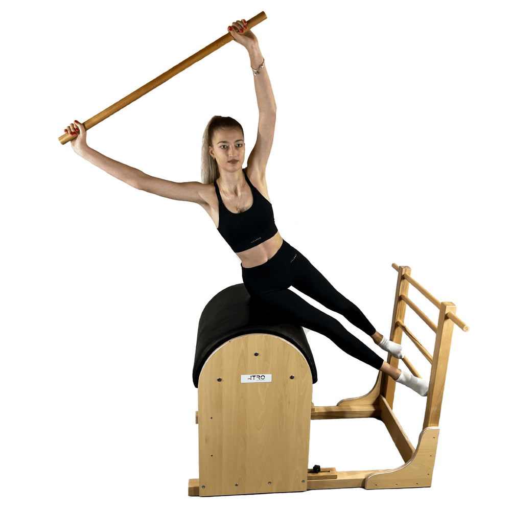 A woman performs a pilates exercise on a wooden reformer machine, holding a wooden bar overhead against a solid white background.