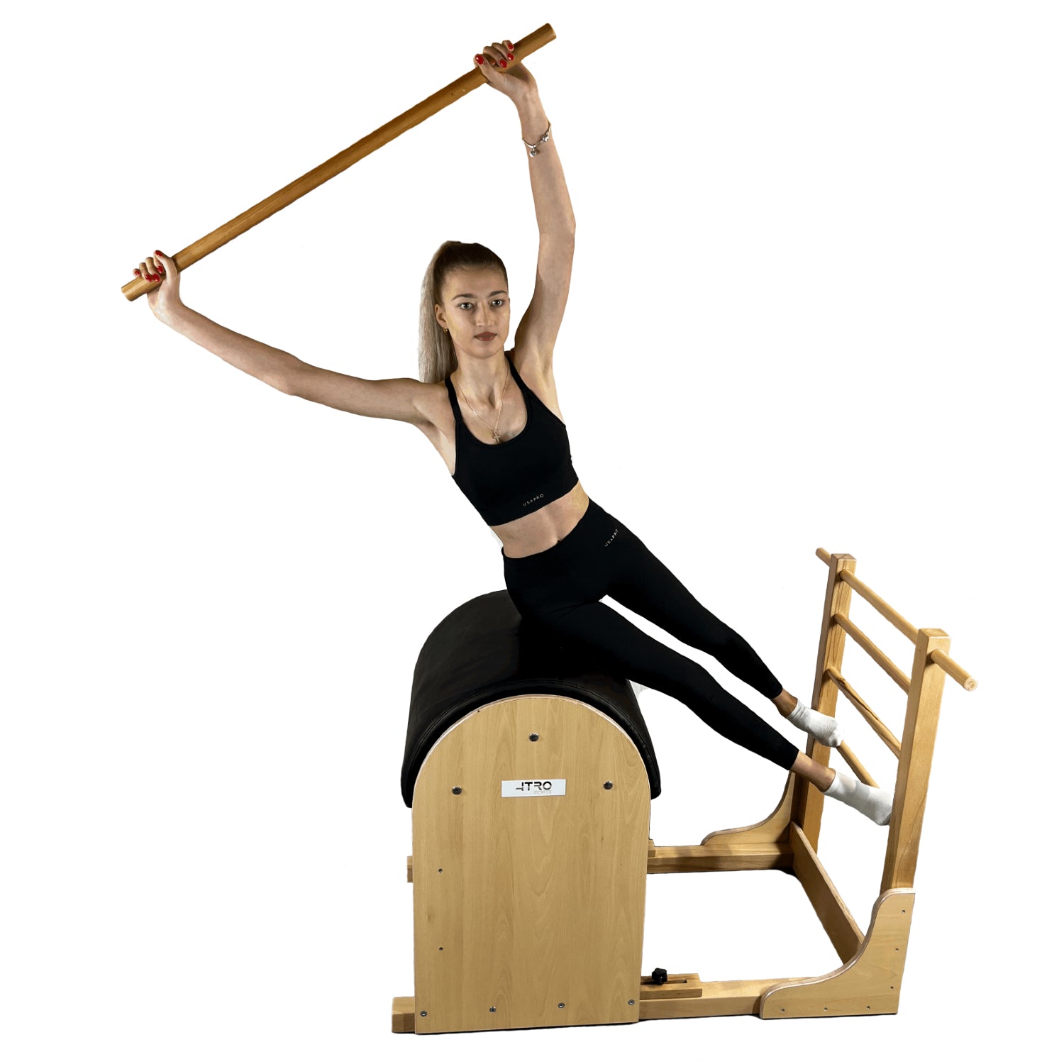 A woman performs a pilates exercise on a wooden reformer machine, holding a wooden bar overhead against a solid white background.